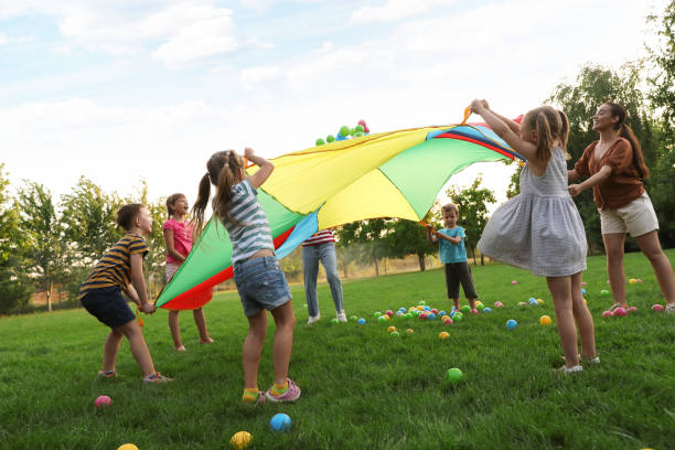 Group of children and teacher playing with rainbow playground parachute on green grass. Summer camp activity